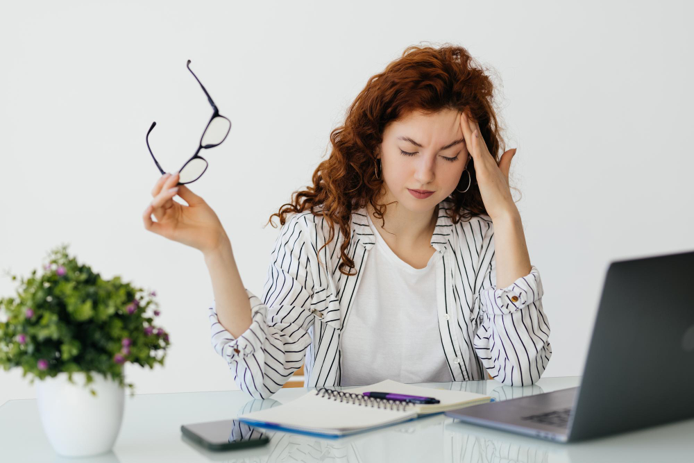 Mulher estressada em frente ao computador e segurando um óculos de grau.