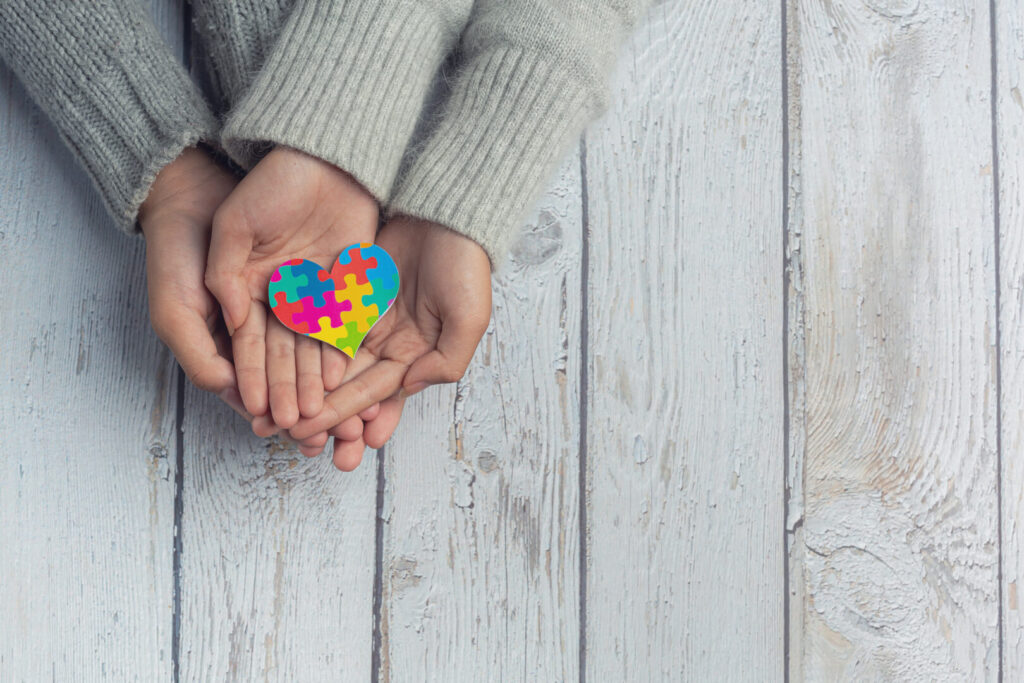 Duas mãos segurando um coração formato por peças de quebra-cabeças coloridas em apoio ao autismo.