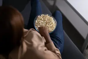 Pessoa sentada no sofá, vista por cima, comendo pipoca em uma tigela grande no colo, representando o ato de assistir a filmes de terror e saúde mental em um ambiente controlado e seguro.