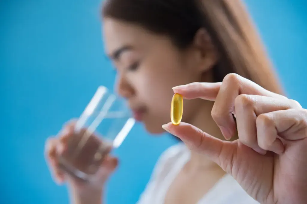 Close-up de uma mão feminina segurando uma cápsula amarela de suplemento de Vitamina B12 (cobalamina) contra um fundo azul, com uma pessoa bebendo água ao fundo, representando o ato de suplementação.