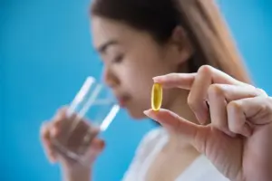Close-up de uma mão feminina segurando uma cápsula amarela de suplemento de Vitamina B12 (cobalamina) contra um fundo azul, com uma pessoa bebendo água ao fundo, representando o ato de suplementação.