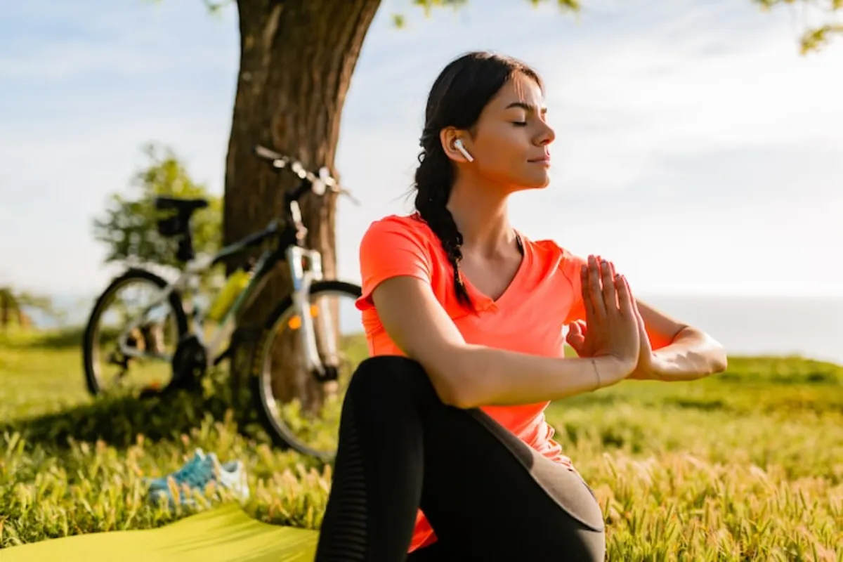 Mulher sentada sobre um tapete ao ar livre, com as mãos em posição de meditação, usando roupa esportiva, com uma bicicleta ao fundo.